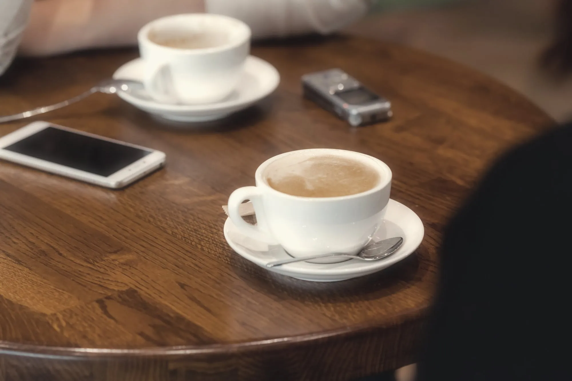 Two cups of coffee on a round wooden café table, with a smartphone and a small voice recorder lying between them.